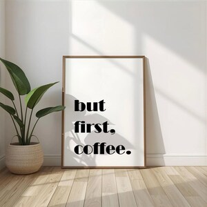 Könnte beinhalten: Ein gerahmter Druck mit den Worten "but first, coffee." in fetter, schwarzer Schrift. Der Druck befindet sich in einem hellbraunen Rahmen und lehnt an einer weißen Wand. Links steht eine Topfpflanze in einem geflochtenen Korb.