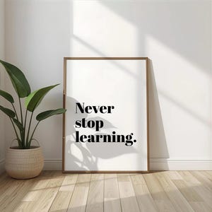 Könnte beinhalten: Gerahmter Druck mit den Worten "Never stop learning" in schwarzer Schrift. Der Druck hat einen Holzrahmen und lehnt an einer weißen Wand. Links vom Druck steht eine Topfpflanze mit großen grünen Blättern.