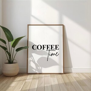 Könnte beinhalten: Ein gerahmter Druck mit den Worten "COFFEE time" in schwarzer Schrift. Der Druck befindet sich in einem hellbraunen Rahmen und lehnt an einer weißen Wand. Links vom Druck steht eine Topfpflanze mit großen grünen Blättern in einem geflochtenen Korb.