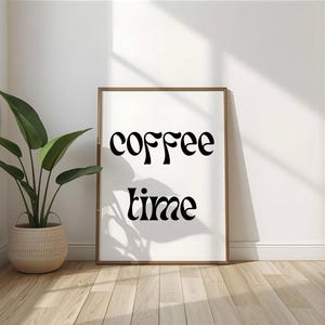 Könnte beinhalten: Ein gerahmter Druck mit den Worten "coffee time" in schwarzer Schrift. Der Druck befindet sich in einem braunen Rahmen und lehnt an einer weißen Wand. Links vom Rahmen steht eine Topfpflanze mit großen grünen Blättern in einem geflochtenen Korb.