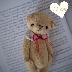 May include: A small, tan teddy bear with a white collar and red ribbon, measuring 4.3 inches tall. The bear has a stitched face and is set against a backdrop of an open book. The text "PDF PATTERN" is visible.