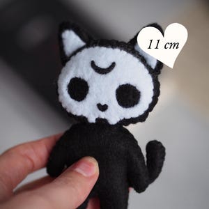 May include: A black and white felt cat doll measuring 11 cm in height. The cat has a white face with black eye sockets, a small heart-shaped nose, and a crescent moon symbol on its forehead. The text PDF PATTERN & INSTRUCTION is visible.