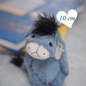 Puede incluir: Un peluche de burro azul, de unos 10 cm de altura, con una melena y cola negras, y un hocico blanco. El juguete tiene orejas rosas y está delante de libros borrosos. El texto "PDF PATTERN" se muestra debajo.