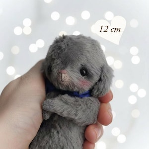 Op de afbeelding: Een grijze pluche konijnenspeelgoed, ongeveer 12 cm hoog, met een blauwe kraag en roze wangen. Het speelgoed wordt in een hand gehouden tegen een vage achtergrond met bokeh-lichten. De tekst "PDF PATTERN" is zichtbaar.
