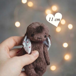 Puede incluir: Un pequeño juguete de peluche de conejo marrón, de aproximadamente 11 cm de altura, con orejas caídas con un patrón de corazón rojo. El juguete tiene una cinta roja alrededor del cuello y se sostiene en una mano. El texto en la imagen dice "PDF PATTERN & INSTRUCTIONS".