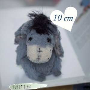May include: A plush, gray donkey toy with a black mane and a white face, approximately 4 inches tall. The toy is sitting on a book, with the text "PDF PATTERN" visible. The donkey has a stitched cross on its face.