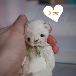 Puede incluir: Un pequeño oso de peluche blanco con una cinta verde alrededor del cuello. El oso mide aproximadamente 9 cm de alto. El texto "PDF PATTERN" es visible. El oso tiene una expresión dulce y está sostenido en una mano.
