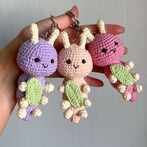 May include: Three handmade crochet bunny keychains in purple, pink, and peach. Each bunny has cream-colored ears and feet, a green leaf-shaped accessory, and a silver keychain ring. The bunnies have black eyes and smiling faces.