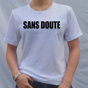 May include: White t-shirt with the phrase "SANS DOUTE" printed in bold black letters across the chest. The shirt has short sleeves and a crew neck. A person is wearing the shirt, with a watch on their wrist.
