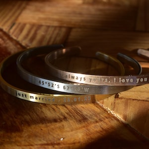 May include: Three engraved metal cuff bracelets in gold, silver, and copper tones. The bracelets have text that reads "just married", "45°52'S 67°30'W", and "always yours, I love you". The bracelets are on a wooden surface.