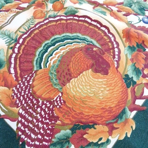 May include: A fabric tablecloth featuring a central illustration of a turkey with a fanned tail in shades of red, orange, and green. The turkey is surrounded by a border of autumn leaves, vines, and berries on a white background with a dark green edge.