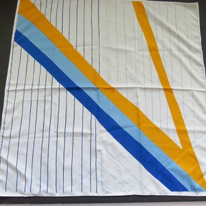 May include: A white silk scarf with a geometric design featuring blue, yellow, and light blue stripes. The scarf has a white background with thin blue vertical stripes and a diagonal stripe pattern in blue, light blue, and yellow. The scarf is signed "Usma" in the bottom right corner.