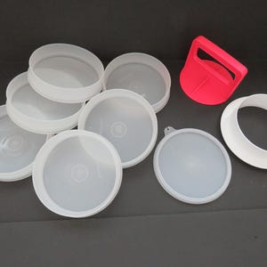 May include: An assortment of white plastic lids and a red handle. The lids are circular with a raised edge. The handle is a bright red color and has a curved shape. The items are arranged on a dark surface.