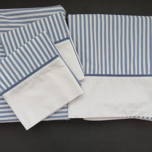 May include: A bedding set featuring a flat sheet, fitted sheet, and two pillowcases. The sheets have a blue and white striped pattern with a white border. The pillowcases have a white border with a blue trim. The set is made of a soft, comfortable material.