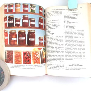 May include: An open cookbook page featuring color photographs of glass jars filled with preserved fruits and vegetables on shelves, alongside printed recipes for olive oil pickles, curry pickles, and bread-and-butter pickles with text.