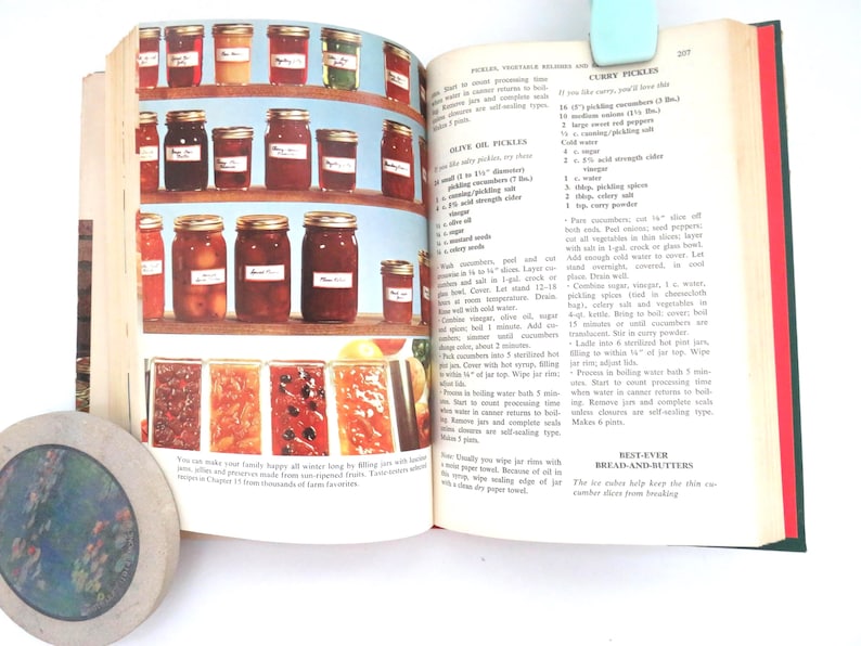 May include: An open cookbook page featuring color photographs of glass jars filled with preserved fruits and vegetables on shelves, alongside printed recipes for olive oil pickles, curry pickles, and bread-and-butter pickles with text.