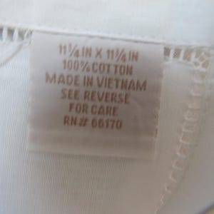 May include: Close-up of a white textile with a tag. The tag reads "11 1/4 in x 11 1/4 in, 100% Cotton, Made in Vietnam, See Reverse for Care, RN# 66170". The textile has a delicate, openwork border.