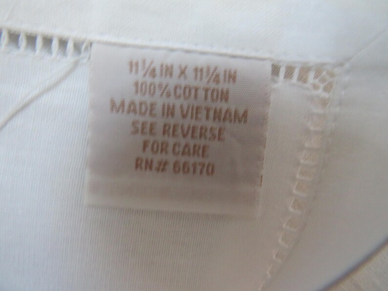May include: Close-up of a white textile with a tag. The tag reads "11 1/4 in x 11 1/4 in, 100% Cotton, Made in Vietnam, See Reverse for Care, RN# 66170". The textile has a delicate, openwork border.