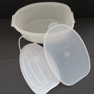 Op de afbeelding: Een set heldere, ovaalvormige Tupperware-containers. De set bevat een grote, open container met handvatten, een deksel en een kleinere, geribbelde lade met een handvat. Het Tupperware-logo is zichtbaar op het deksel.