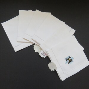 May include: A stack of white linen napkins with a decorative embroidered design in the corner. The napkins are neatly folded and arranged on a dark surface. Each napkin has a tag attached.