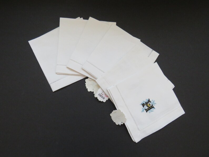 May include: A stack of white linen napkins with a decorative embroidered design in the corner. The napkins are neatly folded and arranged on a dark surface. Each napkin has a tag attached.