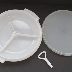 Puede incluir: Bandeja de servir de plástico blanco con tres secciones divididas y una tapa. La bandeja tiene un asa a cada lado. La tapa tiene un pequeño pomo de plástico blanco en el centro.