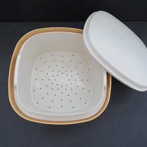 Puede incluir: Un recipiente de plástico blanco y beige para alimentos con tapa. El recipiente tiene forma cuadrada con esquinas redondeadas y una bandeja interior perforada. La tapa está a un lado.