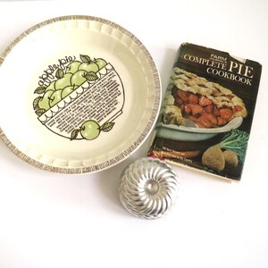 Puede incluir: Un plato para tartas vintage con un dise&ntilde;o de tarta de manzana verde y blanca y las palabras "Apple Pie" impresas en &eacute;l. El plato est&aacute; al lado de un libro de cocina titulado "Farm Fresh Complete Pie Cookbook".