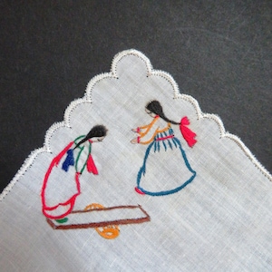 Może przedstawiać: Biała chusteczka z falistymi krawędziami, haftowana dwoma postaciami w kolorowych tradycyjnych strojach. Postacie stoją naprzeciwko siebie przy brązowym stole z pomarańczowym przedmiotem. Haft wykorzystuje czerwoną, niebieską, zieloną i pomarańczową nić.