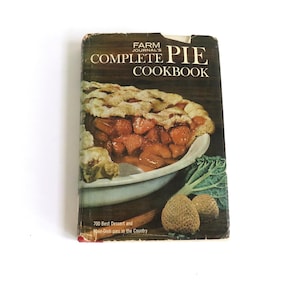 May include: A vintage cookbook titled 'Farm Journal's Complete Pie Cookbook' with a photo of a pie on the cover. The pie is in a white pie dish and has a golden brown crust with a red fruit filling. The background of the cover is brown with green leafy vegetables.