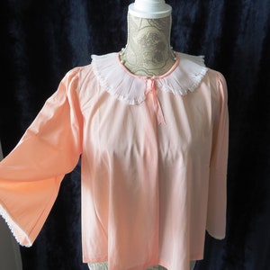 Könnte beinhalten: Eine langärmelige Bluse in Pfirsichfarbe mit einem weißen Spitzenkragen und einer Schleife am Ausschnitt. Die Bluse ist aus einem weichen, seidenartigen Stoff gefertigt.