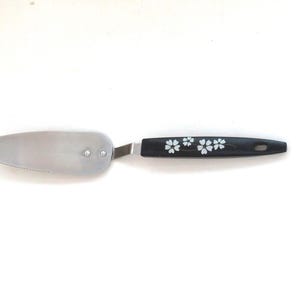 May include: A stainless steel cake server with a serrated edge and a black handle decorated with white floral designs. The server is designed for cutting and serving cakes and pastries.