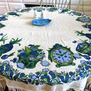 May include: A round, off-white tablecloth with a fringed edge, featuring a repeating border pattern of blue and green clocks, fruit bowls, and floral motifs. Two small white salt and pepper shakers and a blue decorative dish sit in the center.