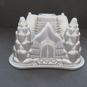 May include: Silver-colored cake pan in the shape of a house with a chimney and two tree-shaped molds on either side. The house has a door and flower details. The pan is made of metal.