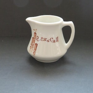 以下が含まれることがあります： 側面に木製の杭の茶色のグラフィックと「Ports o' Call」の文字がプリントされた、白い陶器製のクリーマーピッチャー。ピッチャーには湾曲したハンドルと小さな注ぎ口があり、全体が滑らかで光沢のある仕上がりになっています。