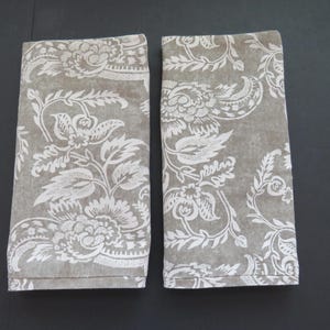May include: Two rectangular cloth napkins with a floral pattern. The napkins are a muted gray color with a white floral design. The floral design includes large flowers and leaves. The napkins are folded and displayed side by side.