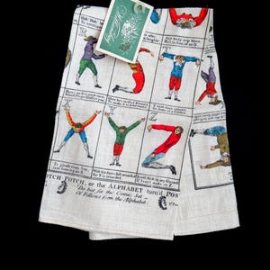 Puede incluir: Paño de cocina de lino blanco con un diseño de alfabeto vintage. El paño presenta figuras con trajes coloridos que forman letras, con texto adjunto. Se adjunta una etiqueta que dice "Williamtay". El diseño recuerda a una impresión antigua.