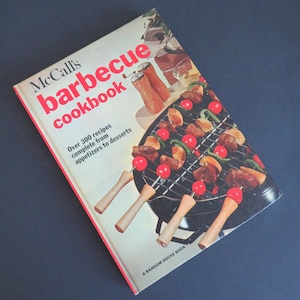 Puede incluir: Un libro de cocina rojo y blanco titulado "McCall's barbecue cookbook" con una foto de comida a la parrilla en brochetas. El texto en la portada dice "Más de 300 recetas completas, desde aperitivos hasta postres".