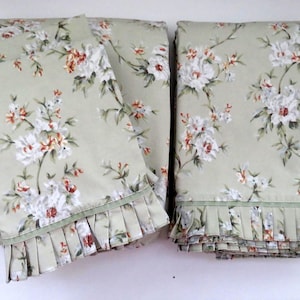 May include: A set of sage green curtains featuring a floral pattern with white and orange blossoms and green leaves. The curtains have a pleated ruffle trim along the bottom edge, adding texture to the fabric. The design is consistent across both panels.