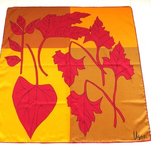 Peut inclure: Un foulard carré en soie présentant un fond color-block composé de quadrants jaune, orange et marron. Le foulard est orné de grandes illustrations de feuilles rouges audacieuses avec des contours noirs. La signature "Usna." est imprimée en noir.