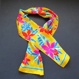 May include: A vibrant yellow scarf with a colorful floral pattern. The scarf features large pink, red, and blue flowers and leaves against a bright yellow background. The scarf is made of a silky material and is a stylish accessory.