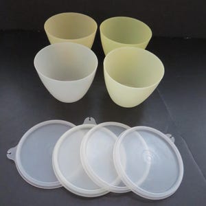 May include: Four light yellow and white plastic bowls with matching clear lids. The bowls are cone-shaped and appear to be made of a durable plastic material. The lids are round and have a small tab for easy removal.