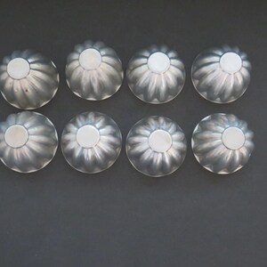 May include: Eight small, silver-colored, fluted molds arranged in two rows of four. Each mold has a circular, raised center. The molds are likely for baking or creating decorative food items. The background is a dark gray.