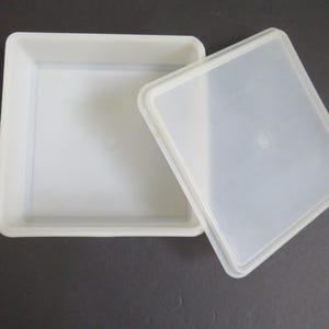 May include: A square, off-white plastic storage container with a matching, slightly translucent, removable lid. The container features a flat base and straight, vertical sides, while the lid is designed to fit securely over the top opening.