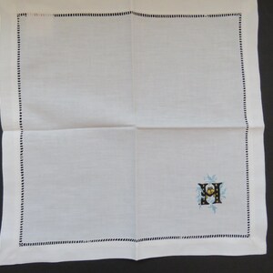 May include: White linen handkerchief with a black stitched border. The handkerchief features an embroidered initial with blue and yellow floral accents in the corner. A classic and elegant accessory.