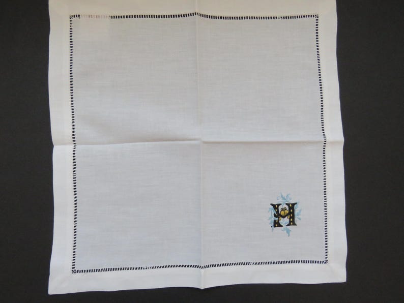 May include: White linen handkerchief with a black stitched border. The handkerchief features an embroidered initial with blue and yellow floral accents in the corner. A classic and elegant accessory.