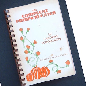 Puede incluir: Un libro de cocina encuadernado en espiral titulado "The Compleat Pumpkin Eater" con letras naranjas e ilustraciones de calabazas. La portada es blanquecina con una encuadernación en espiral marrón.