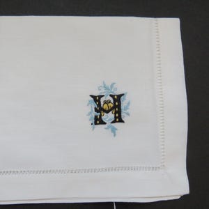 May include: White linen napkin with a decorative embroidered letter "H". The letter is black with yellow and blue accents, surrounded by a light blue floral design. The napkin has a stitched border.