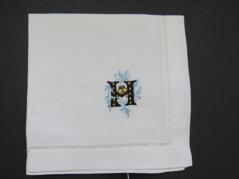 May include: White linen napkin with a decorative embroidered letter "H". The letter is black with yellow and blue accents, surrounded by a light blue floral design. The napkin has a stitched border.