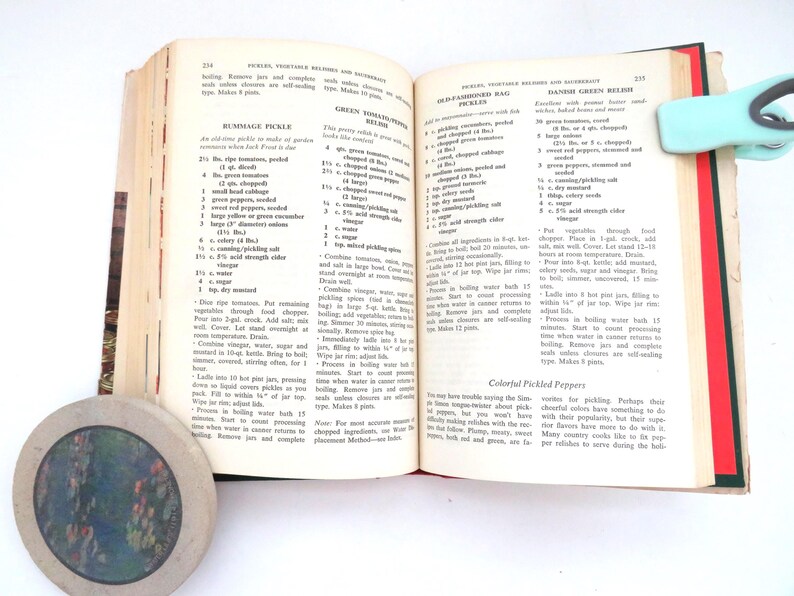 May include: An open cookbook with pages 234 and 235 visible, featuring recipes for Rummage Pickle, Green Tomato Relish, Old-Fashioned Rag Pickles, and Danish Green Relish. A light blue clip holds the top right corner, and a round coaster is nearby.